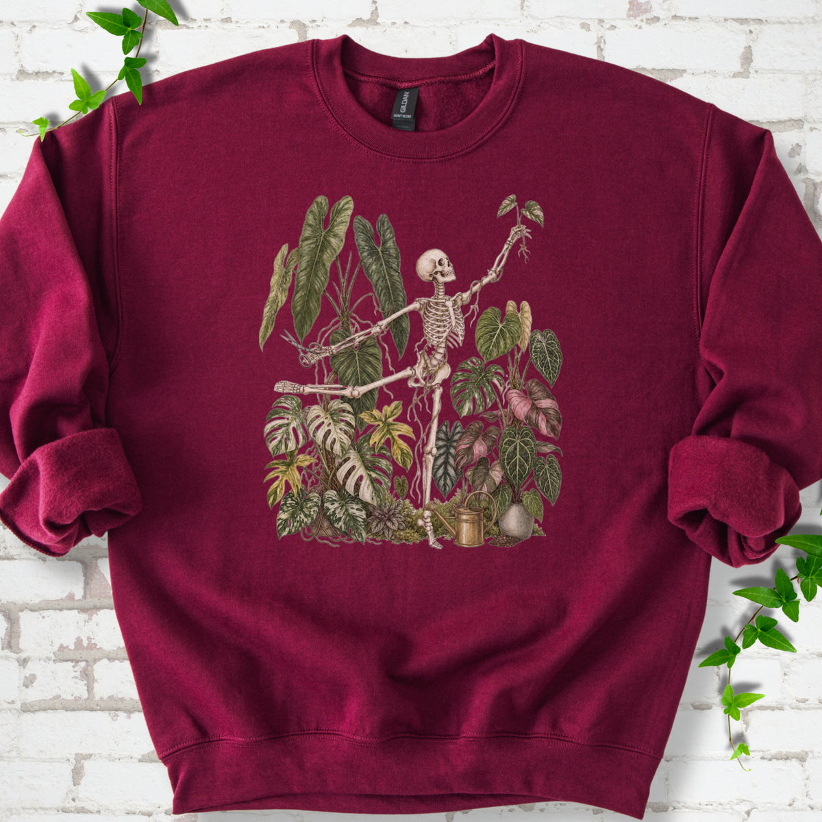 Maroon Gildan® 18000 crewneck sweatshirt flat lay featuring a skeleton tending a collection of tropical houseplants.