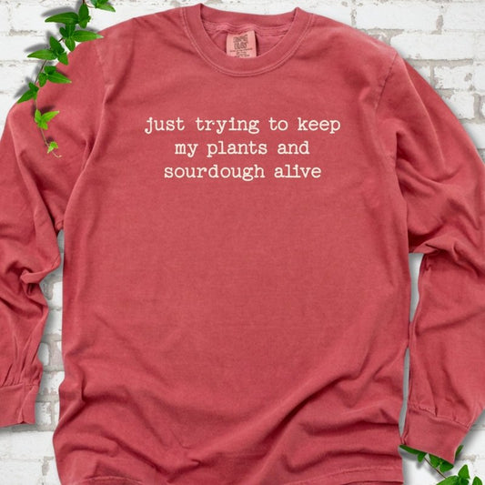 Crimson Comfort Colors® long sleeve T-shirt front mockup featuring the words “just trying to keep my plants and sourdough alive.”