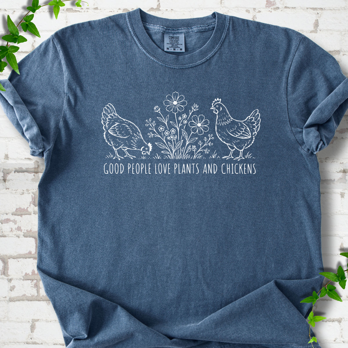 Denim T-shirt flat lay with white line-art hens and flowers above the words "Good People Love Plants and Chickens."