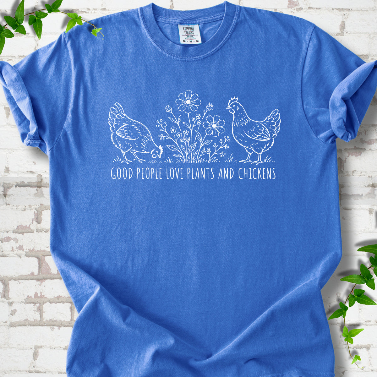 Flo Blue T-shirt flat lay with white line-art hens and flowers above the words "Good People Love Plants and Chickens."