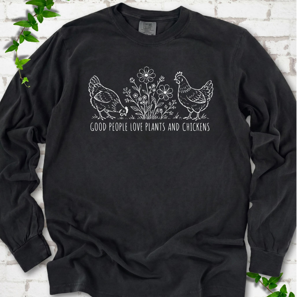 Black long-sleeve T-shirt flat lay with white line-art hens and flowers above the words "Good People Love Plants and Chickens."