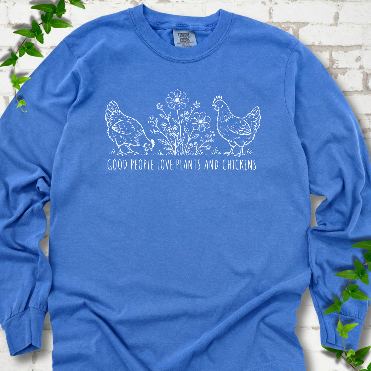 Flo Blue long-sleeve T-shirt flat lay with white line-art hens and flowers above the words "Good People Love Plants and Chickens."