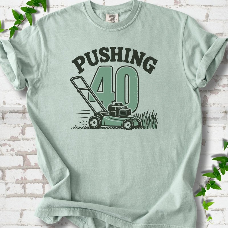 Bay short sleeve T-shirt flat lay with dark arched "PUSHING" lettering above a large green 40 and a push lawn mower in the grass, on white brick with ivy sprigs.