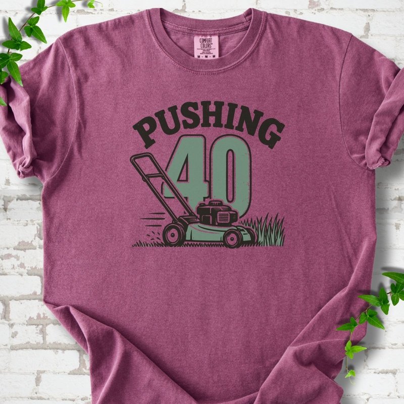 Berry short sleeve T-shirt flat lay with dark arched "PUSHING" lettering above a large green 40 and a push lawn mower in the grass, on white brick with ivy sprigs.