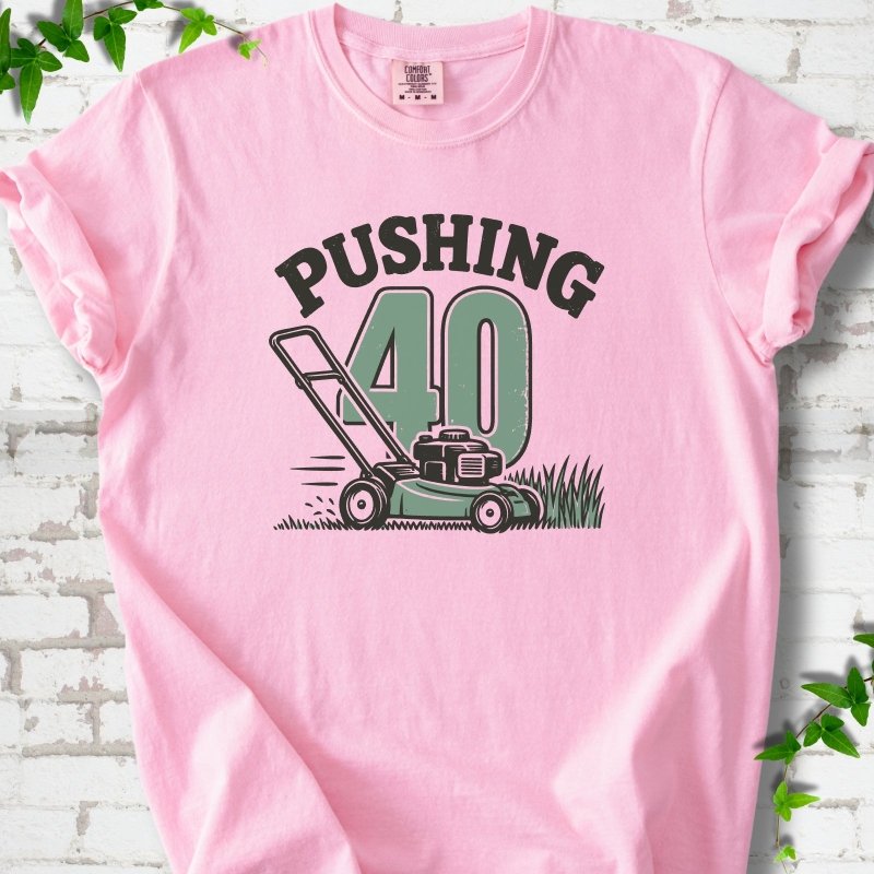 Blossom short sleeve T-shirt flat lay with dark arched "PUSHING" lettering above a large green 40 and a push lawn mower in the grass, on white brick with ivy sprigs.