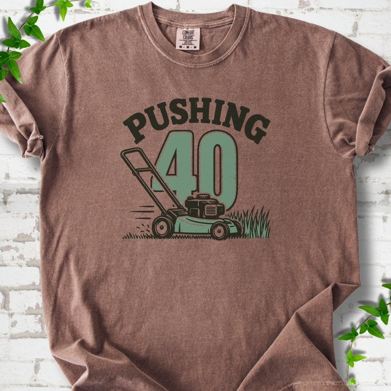 Espresso short sleeve T-shirt flat lay with dark arched "PUSHING" lettering above a large green 40 and a push lawn mower in the grass, on white brick with ivy sprigs.