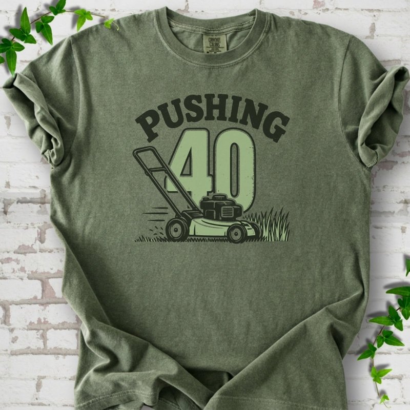 Moss short sleeve T-shirt flat lay with dark arched "PUSHING" lettering above a large green 40 and a push lawn mower in the grass, on white brick with ivy sprigs.