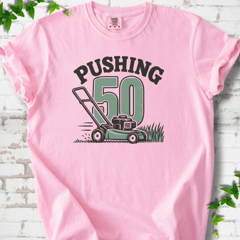 Blossom short sleeve T-shirt flat lay with dark arched "PUSHING" lettering above a large green 50 and a push lawn mower in the grass, on white brick with ivy sprigs.