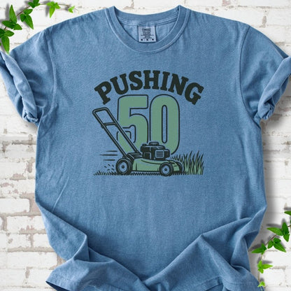 Blue Jean short sleeve T-shirt flat lay with dark arched "PUSHING" lettering above a large green 50 and a push lawn mower in the grass, on white brick with ivy sprigs.
