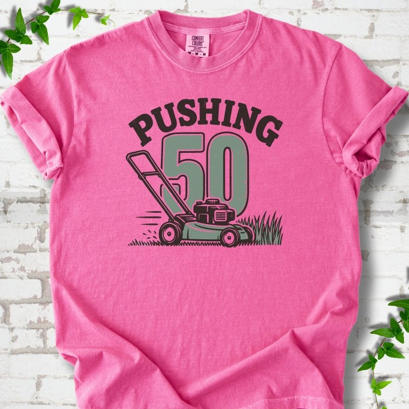 Crunchberry short sleeve T-shirt flat lay with dark arched "PUSHING" lettering above a large green 50 and a push lawn mower in the grass, on white brick with ivy sprigs.