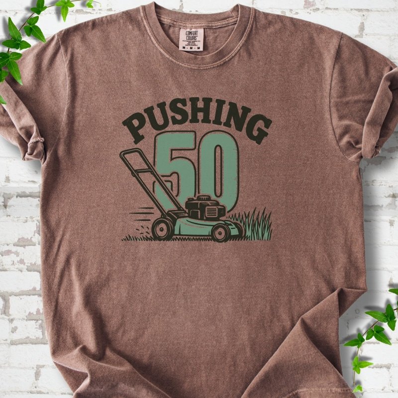 Espresso short sleeve T-shirt flat lay with dark arched "PUSHING" lettering above a large green 50 and a push lawn mower in the grass, on white brick with ivy sprigs.