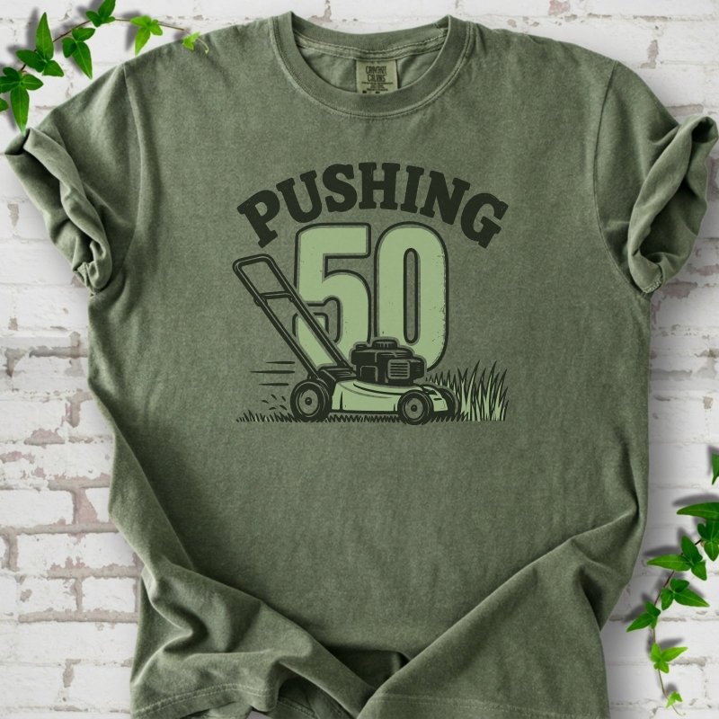 Moss short sleeve T-shirt flat lay with dark arched "PUSHING" lettering above a large green 50 and a push lawn mower in the grass, on white brick with ivy sprigs.