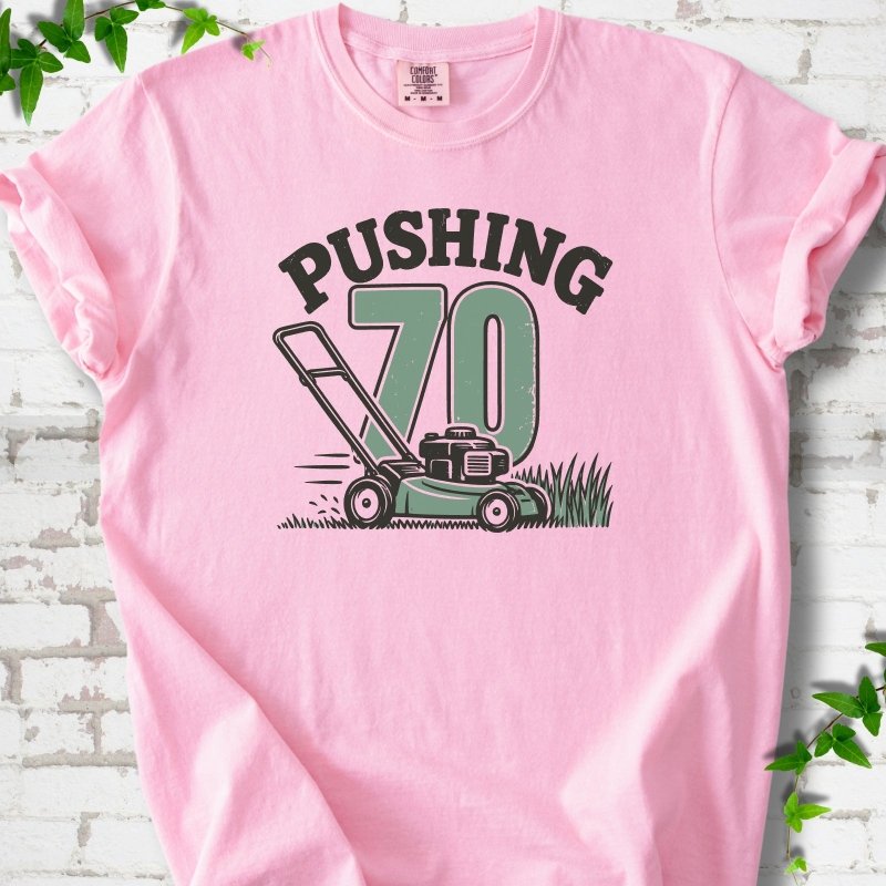 Blossom short sleeve T-shirt flat lay with dark arched "PUSHING" lettering above a large green 70 and a push lawn mower in the grass, on white brick with ivy sprigs.