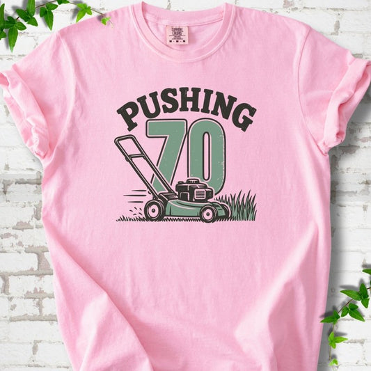 Blossom short sleeve T-shirt flat lay with dark arched "PUSHING" lettering above a large green 70 and a push lawn mower in the grass, on white brick with ivy sprigs.