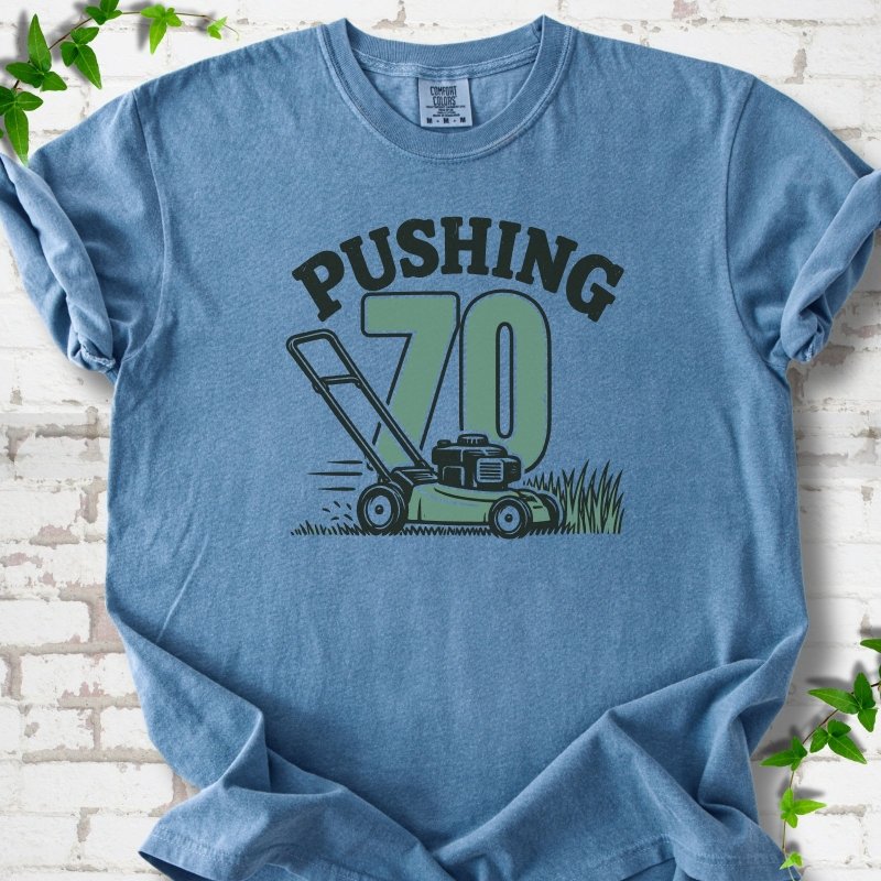 Blue Jean short sleeve T-shirt flat lay with dark arched "PUSHING" lettering above a large green 70 and a push lawn mower in the grass, on white brick with ivy sprigs.
