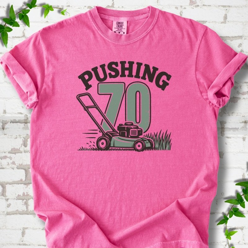 Crunchberry short sleeve T-shirt flat lay with dark arched "PUSHING" lettering above a large green 70 and a push lawn mower in the grass, on white brick with ivy sprigs.