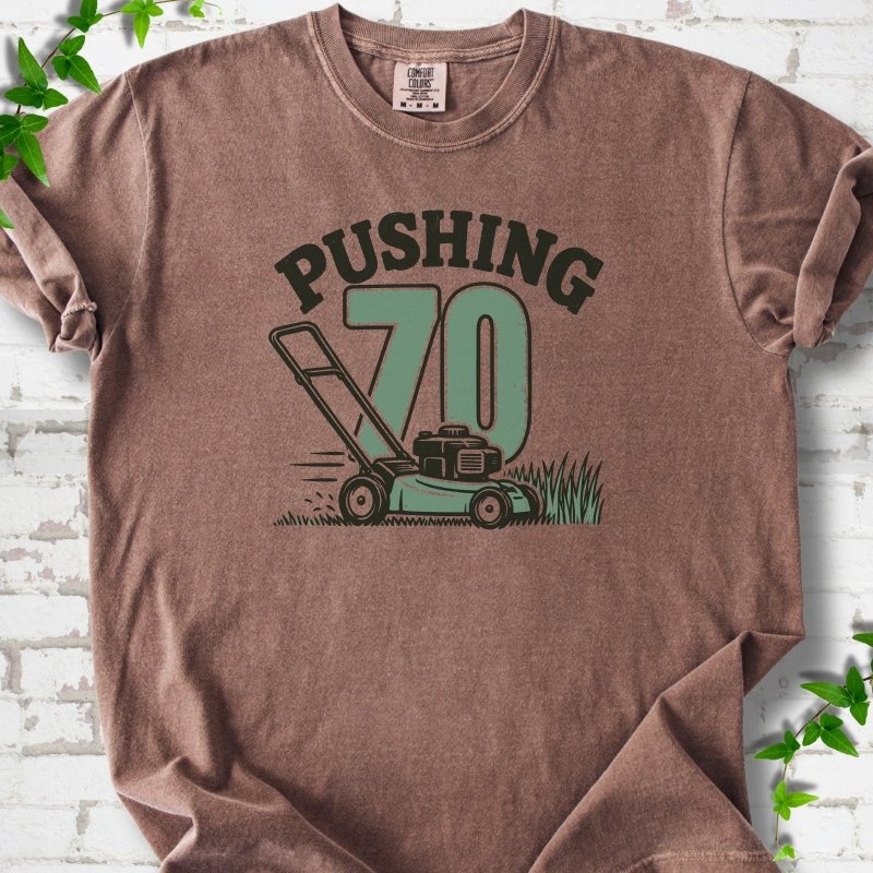 Espresso short sleeve T-shirt flat lay with dark arched "PUSHING" lettering above a large green 70 and a push lawn mower in the grass, on white brick with ivy sprigs.