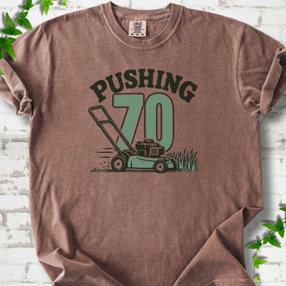 Espresso short sleeve T-shirt flat lay with dark arched "PUSHING" lettering above a large green 70 and a push lawn mower in the grass, on white brick with ivy sprigs.