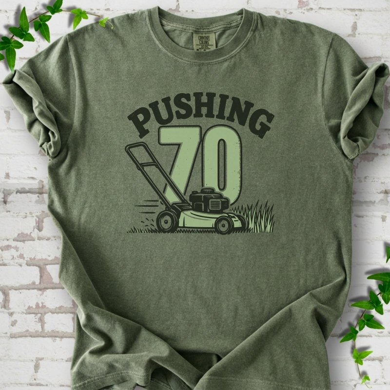Moss short sleeve T-shirt flat lay with dark arched "PUSHING" lettering above a large green 70 and a push lawn mower in the grass, on white brick with ivy sprigs.
