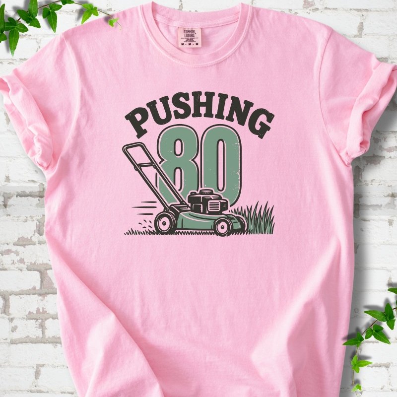 Blossom short sleeve T-shirt flat lay with dark arched "PUSHING" lettering above a large green 80 and a push lawn mower in the grass, on white brick with ivy sprigs.