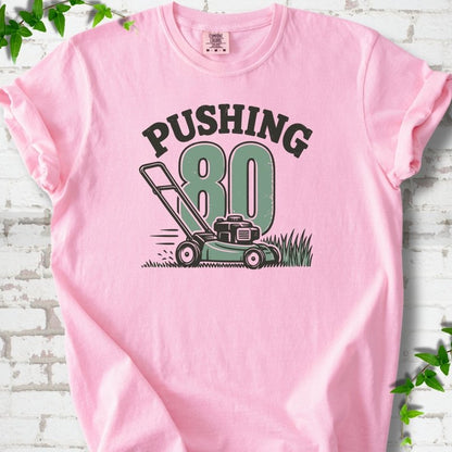 Blossom short sleeve T-shirt flat lay with dark arched "PUSHING" lettering above a large green 80 and a push lawn mower in the grass, on white brick with ivy sprigs.