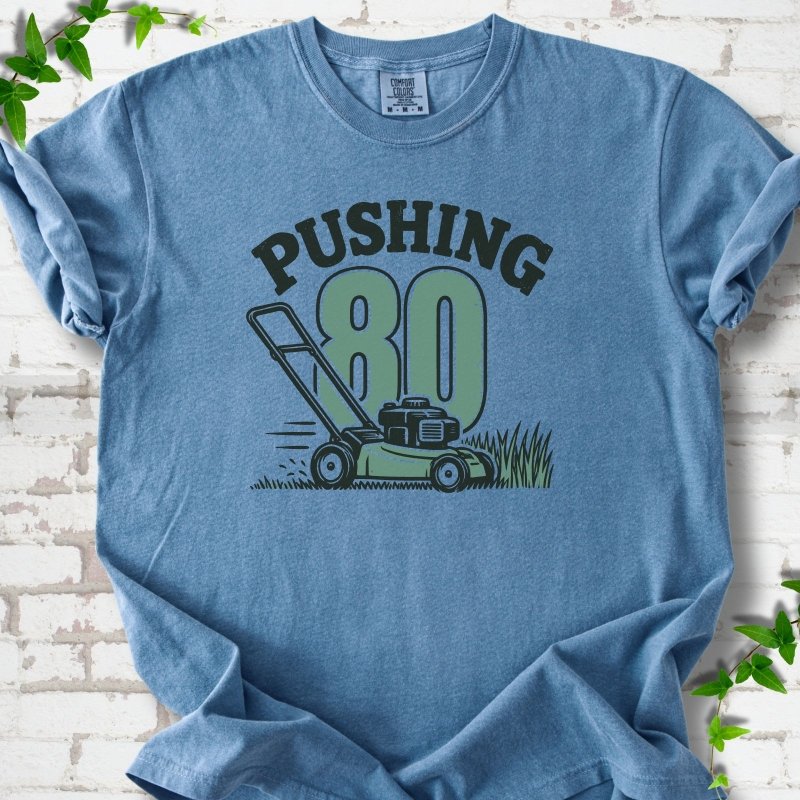 Blue Jean short sleeve T-shirt flat lay with dark arched "PUSHING" lettering above a large green 80 and a push lawn mower in the grass, on white brick with ivy sprigs.