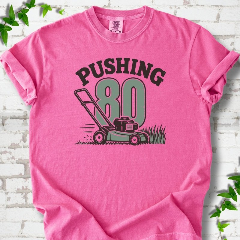 Crunchberry short sleeve T-shirt flat lay with dark arched "PUSHING" lettering above a large green 80 and a push lawn mower in the grass, on white brick with ivy sprigs.