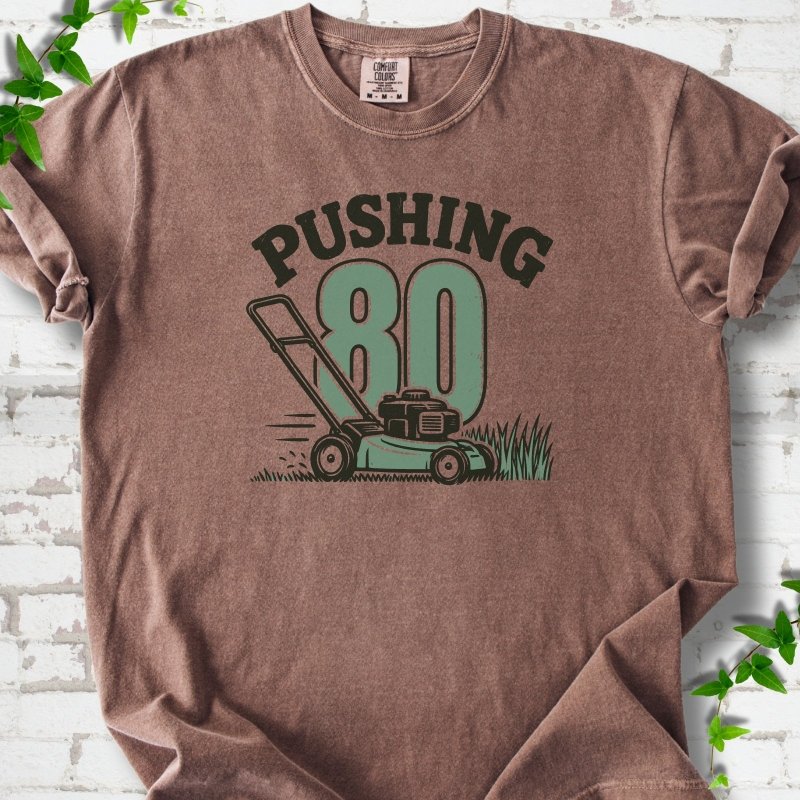 Espresso short sleeve T-shirt flat lay with dark arched "PUSHING" lettering above a large green 80 and a push lawn mower in the grass, on white brick with ivy sprigs.
