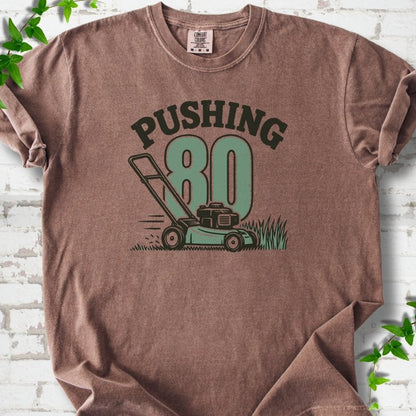 Espresso short sleeve T-shirt flat lay with dark arched "PUSHING" lettering above a large green 80 and a push lawn mower in the grass, on white brick with ivy sprigs.