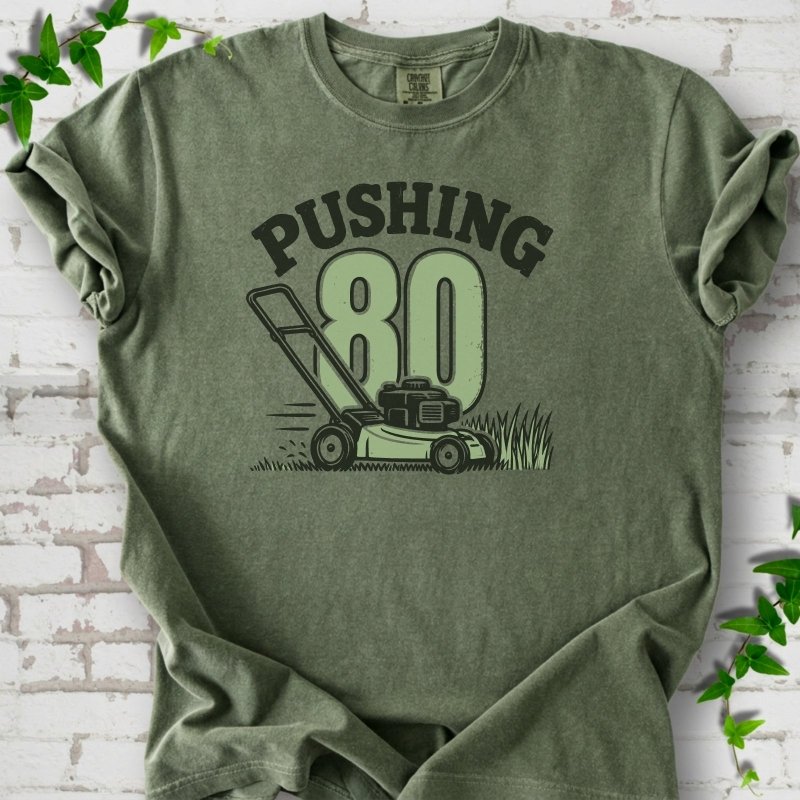 Moss short sleeve T-shirt flat lay with dark arched "PUSHING" lettering above a large green 80 and a push lawn mower in the grass, on white brick with ivy sprigs.