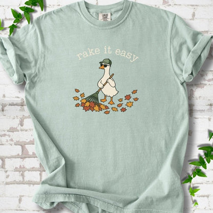 Bay T-shirt flat lay with ivory "rake it easy" lettering above a white goose in a green cap and sunglasses raking a pile of orange and gold fall leaves, on white brick with ivy sprigs.