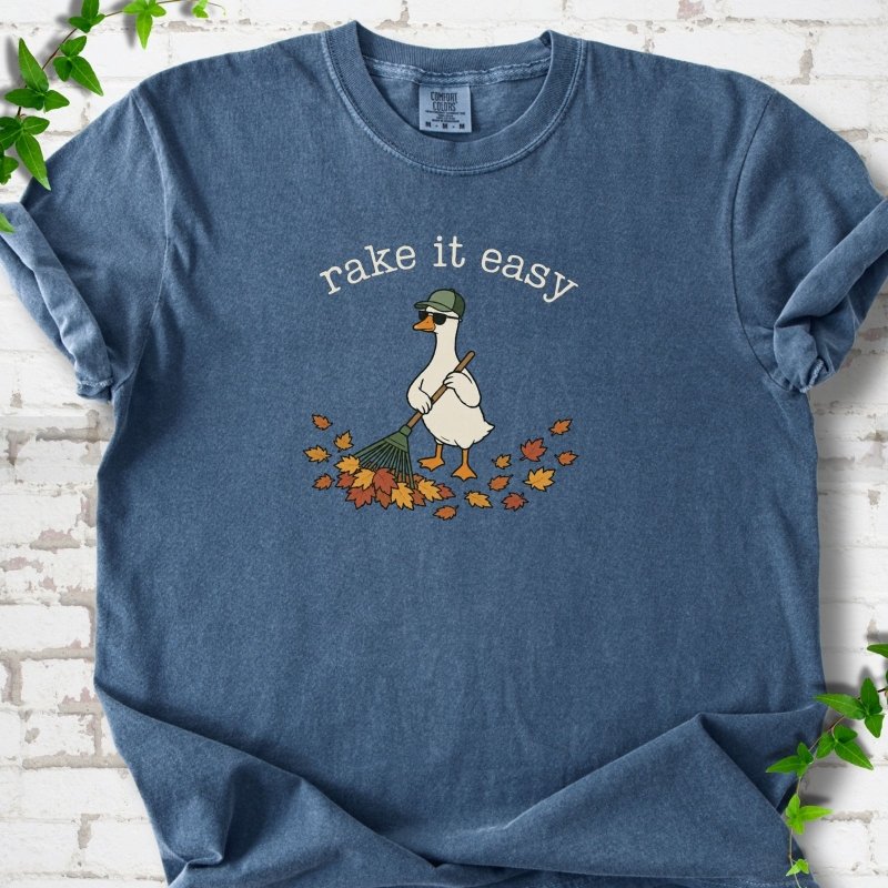 Denim T-shirt flat lay with ivory "rake it easy" lettering above a white goose in a green cap and sunglasses raking a pile of orange and gold fall leaves, on white brick with ivy sprigs.