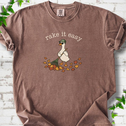 Espresso T-shirt flat lay with ivory "rake it easy" lettering above a white goose in a green cap and sunglasses raking a pile of orange and gold fall leaves, on white brick with ivy sprigs.