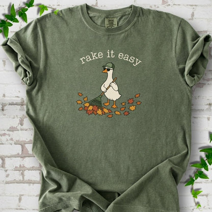 Moss T-shirt flat lay with ivory "rake it easy" lettering above a white goose in a green cap and sunglasses raking a pile of orange and gold fall leaves, on white brick with ivy sprigs.