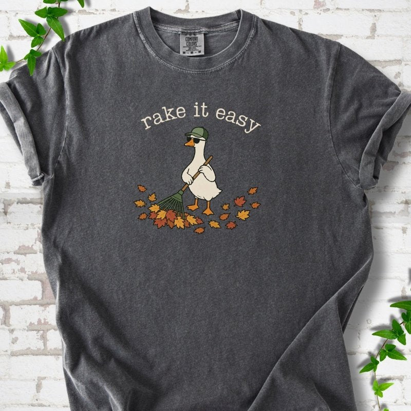 Pepper T-shirt flat lay with ivory "rake it easy" lettering above a white goose in a green cap and sunglasses raking a pile of orange and gold fall leaves, on white brick with ivy sprigs.