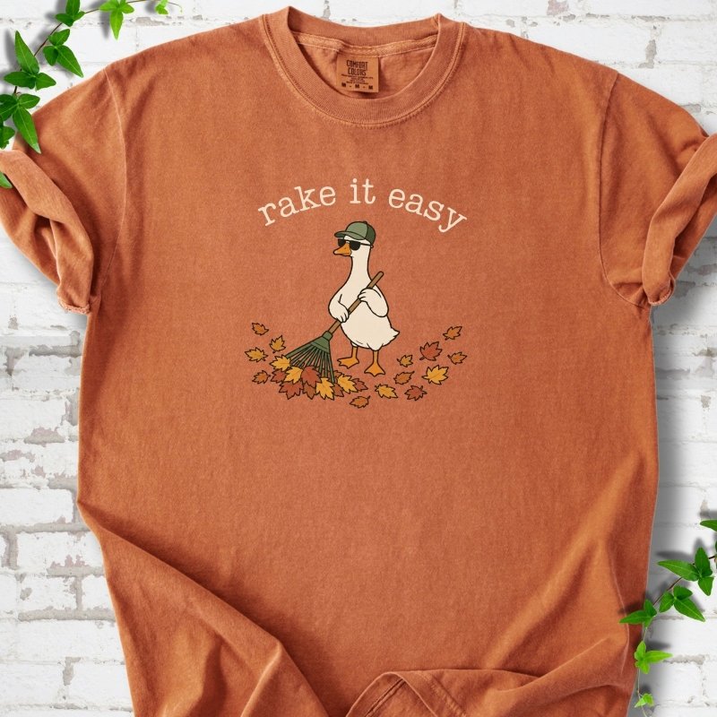 Yam T-shirt flat lay with ivory "rake it easy" lettering above a white goose in a green cap and sunglasses raking a pile of orange and gold fall leaves, on white brick with ivy sprigs.