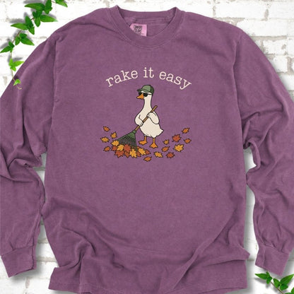 Berry long sleeve T-shirt flat lay with ivory "rake it easy" lettering above a white goose in a green cap and sunglasses raking a pile of orange and gold fall leaves, on white brick with ivy sprigs.