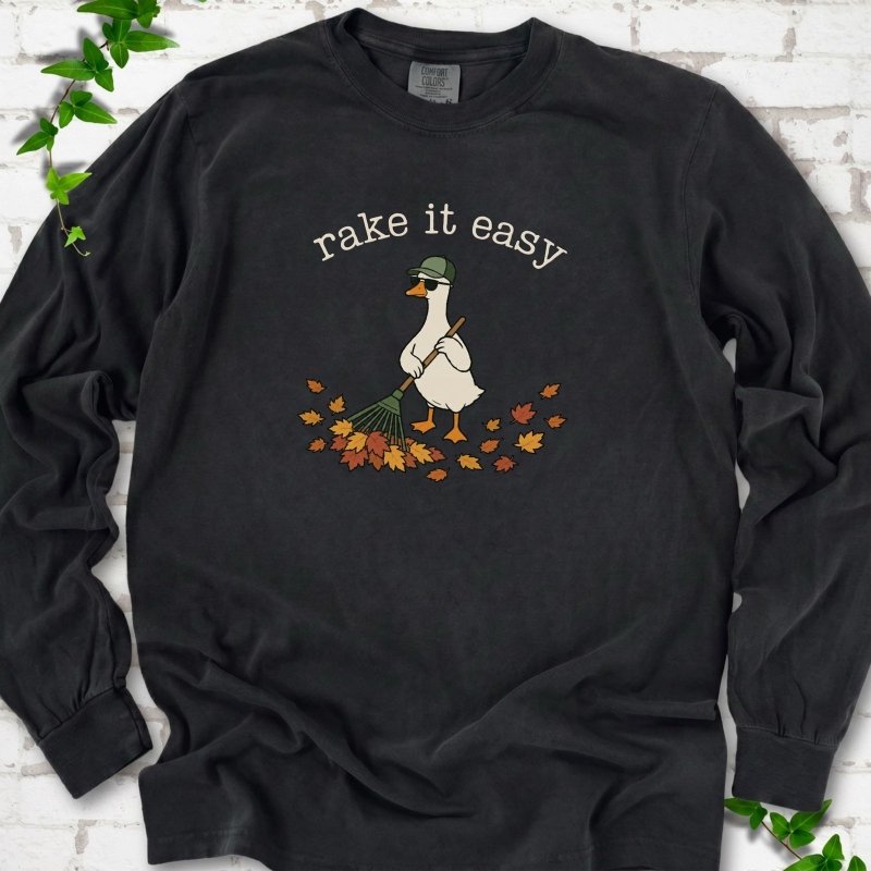Black long sleeve T-shirt flat lay with ivory "rake it easy" lettering above a white goose in a green cap and sunglasses raking a pile of orange and gold fall leaves, on white brick with ivy sprigs.