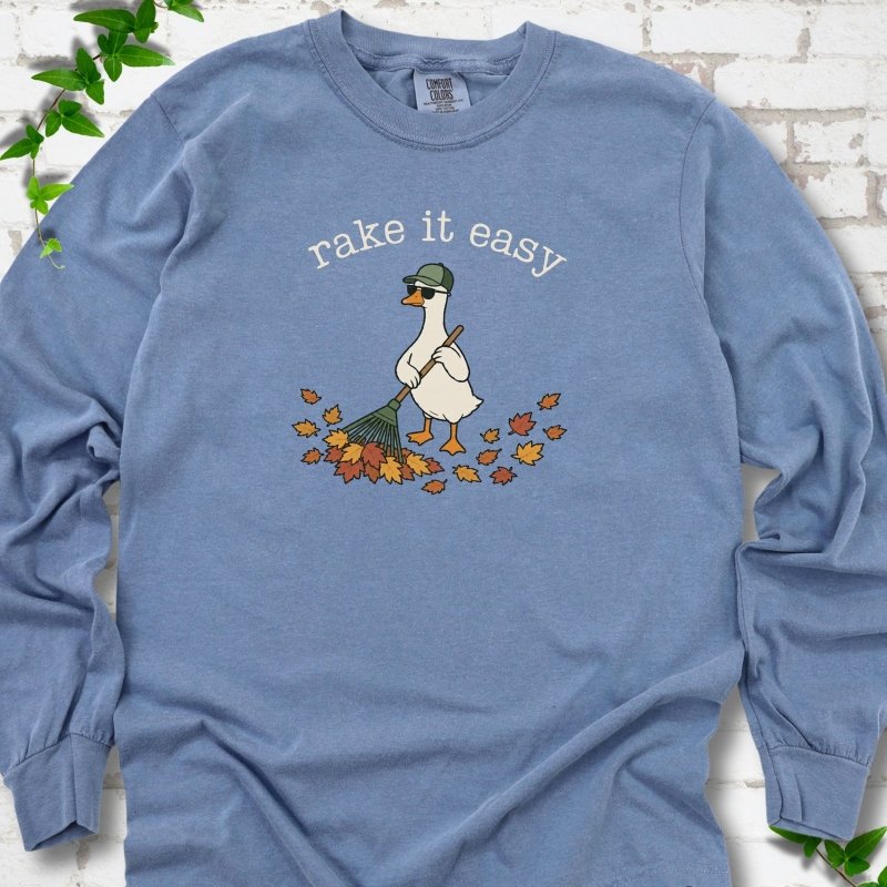 Blue Jean long sleeve T-shirt flat lay with ivory "rake it easy" lettering above a white goose in a green cap and sunglasses raking a pile of orange and gold fall leaves, on white brick with ivy sprigs.