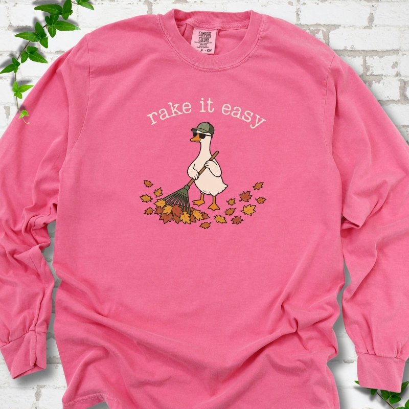 Crunchberry long sleeve T-shirt flat lay with ivory "rake it easy" lettering above a white goose in a green cap and sunglasses raking a pile of orange and gold fall leaves, on white brick with ivy sprigs.