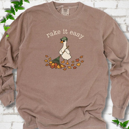 Espresso long sleeve T-shirt flat lay with ivory "rake it easy" lettering above a white goose in a green cap and sunglasses raking a pile of orange and gold fall leaves, on white brick with ivy sprigs.