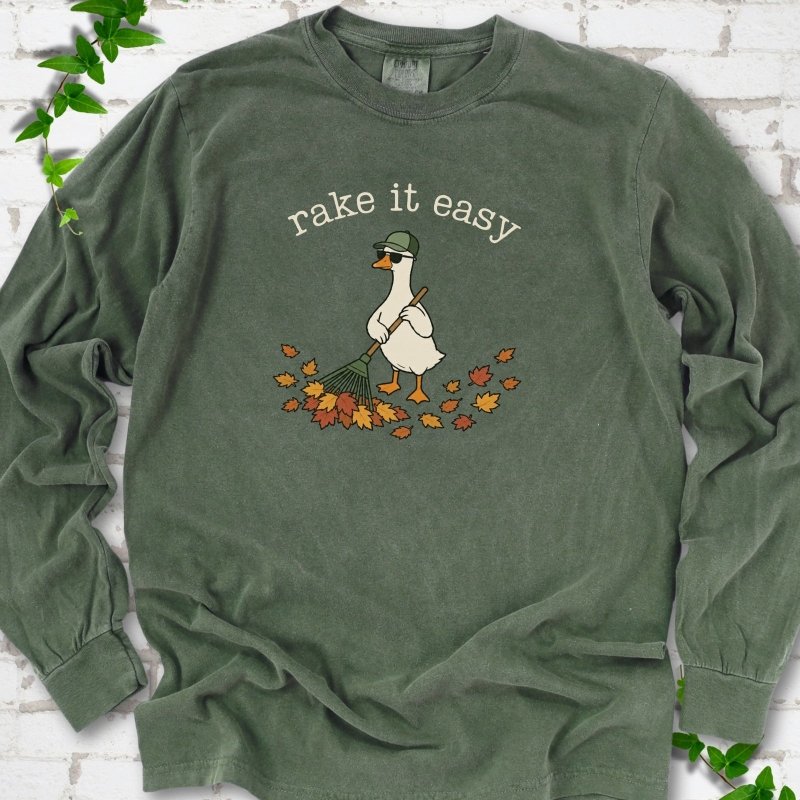 Hemp long sleeve T-shirt flat lay with ivory "rake it easy" lettering above a white goose in a green cap and sunglasses raking a pile of orange and gold fall leaves, on white brick with ivy sprigs.