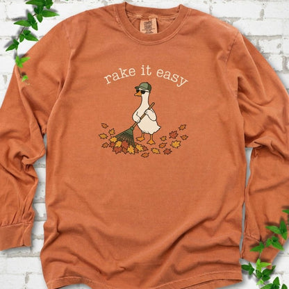 Yam long sleeve T-shirt flat lay with ivory "rake it easy" lettering above a white goose in a green cap and sunglasses raking a pile of orange and gold fall leaves, on white brick with ivy sprigs.