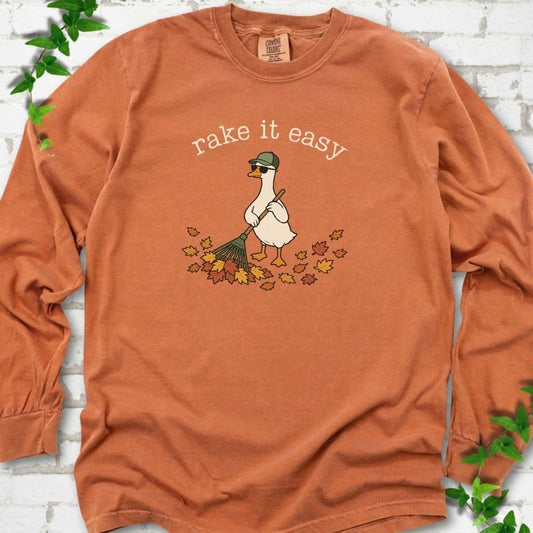 Yam long sleeve T-shirt flat lay with ivory "rake it easy" lettering above a white goose in a green cap and sunglasses raking a pile of orange and gold fall leaves, on white brick with ivy sprigs.