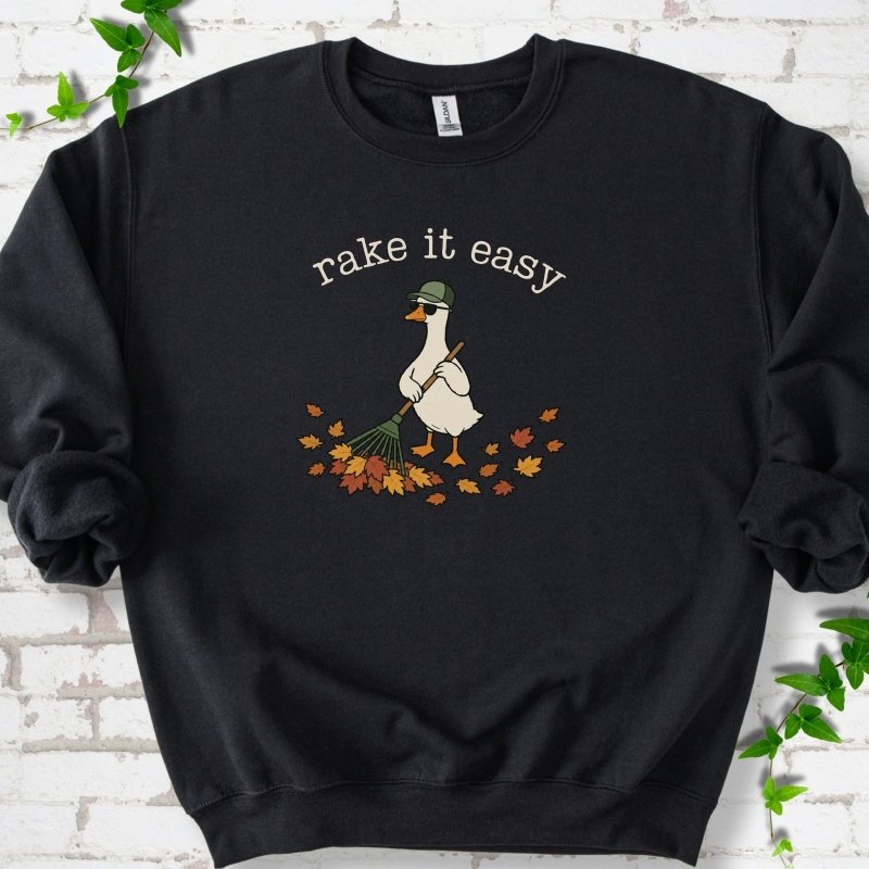Black sweatshirt flat lay with ivory "rake it easy" lettering above a white goose in a green cap and sunglasses raking a pile of orange and gold fall leaves, on white brick with ivy sprigs.