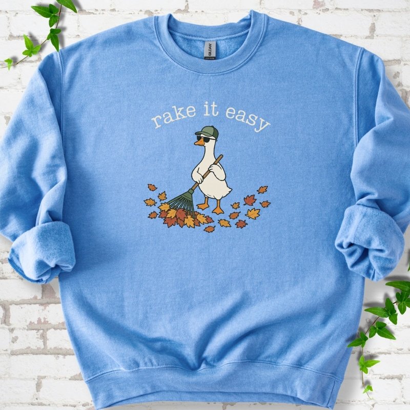 Carolina Blue sweatshirt flat lay with ivory "rake it easy" lettering above a white goose in a green cap and sunglasses raking a pile of orange and gold fall leaves, on white brick with ivy sprigs.