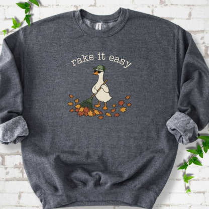 Dark Heather sweatshirt flat lay with ivory "rake it easy" lettering above a white goose in a green cap and sunglasses raking a pile of orange and gold fall leaves, on white brick with ivy sprigs.