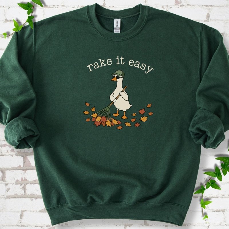 Forest Green sweatshirt flat lay with ivory "rake it easy" lettering above a white goose in a green cap and sunglasses raking a pile of orange and gold fall leaves, on white brick with ivy sprigs.