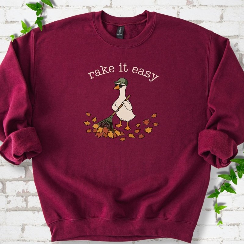 Maroon sweatshirt flat lay with ivory "rake it easy" lettering above a white goose in a green cap and sunglasses raking a pile of orange and gold fall leaves, on white brick with ivy sprigs.
