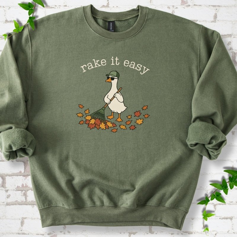 Military Green sweatshirt flat lay with ivory "rake it easy" lettering above a white goose in a green cap and sunglasses raking a pile of orange and gold fall leaves, on white brick with ivy sprigs.